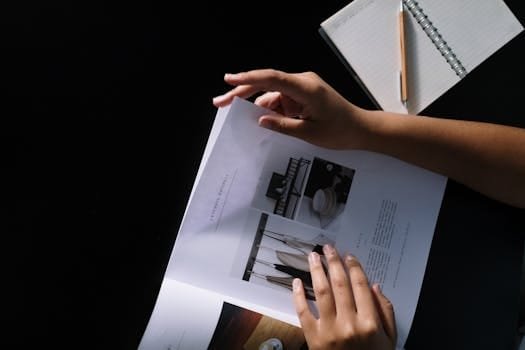 Hands flipping through a design magazine on a dark desk with a notebook and pencil.